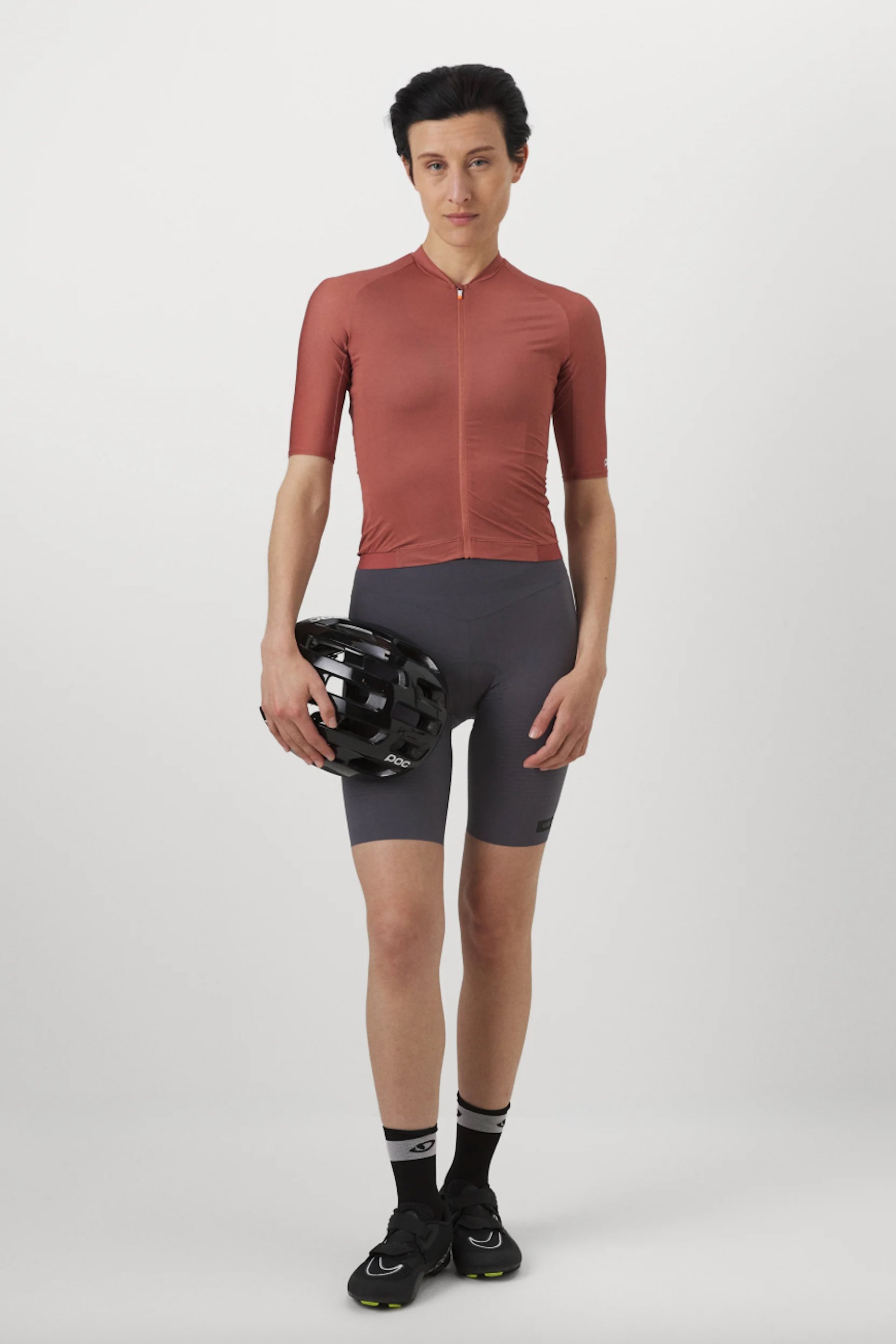 Person in a roten Fahrradjacket und grauen Shorts hält einen schwarzen Fahrradhelm und steht vor einem schlichten weißen Hintergrund.