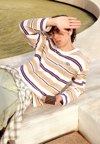 Young man shielding his eyes from the sunlight, wearing a striped jumper and earphones, reclining on a stone ledge near a circular water feature.