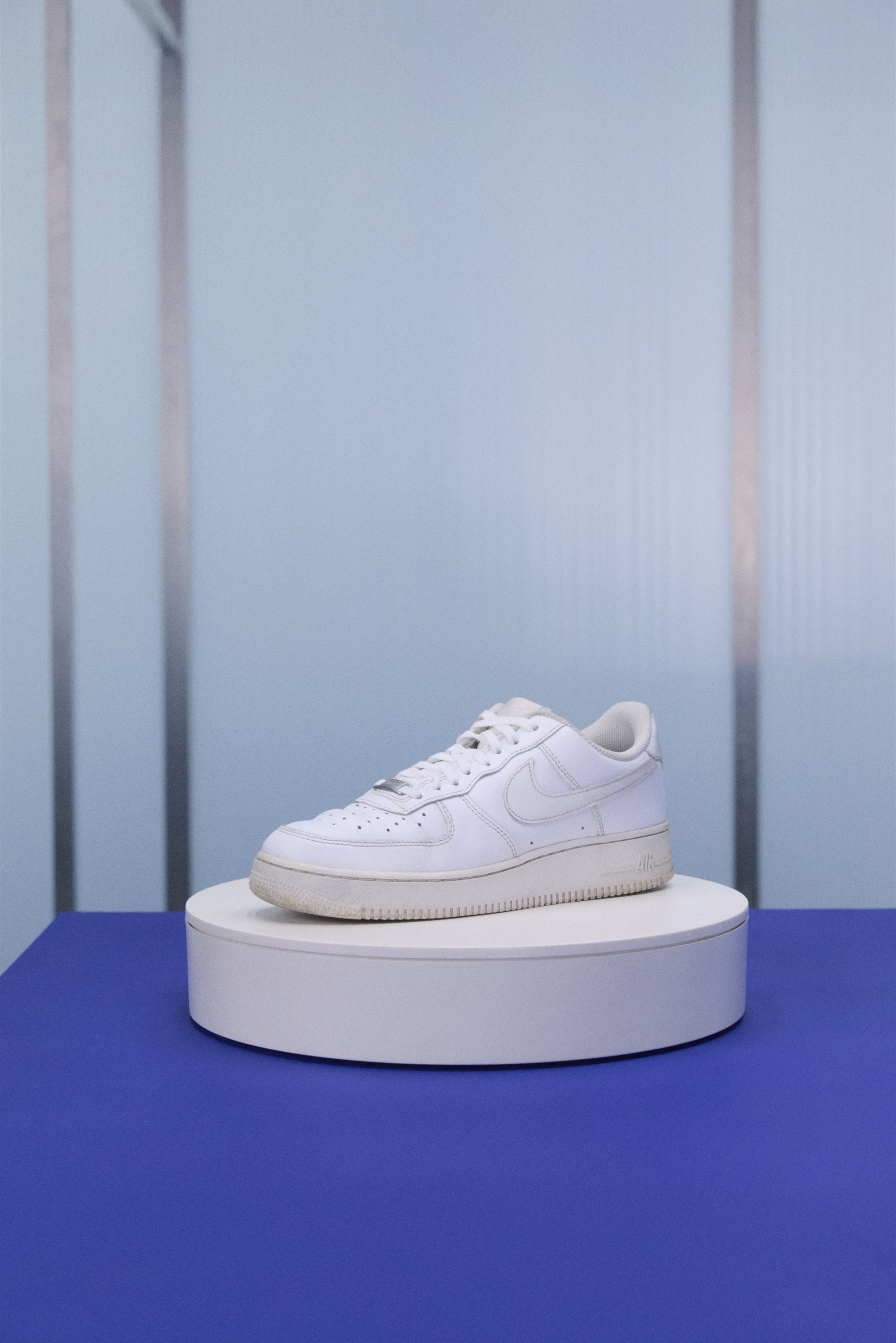 Valkoiset Nike Air Force 1 -kengät esillä valkoisella pyöreällä pylväällä, vaaleansinistä taustaa ja lilaa pöytätasoa vasten.