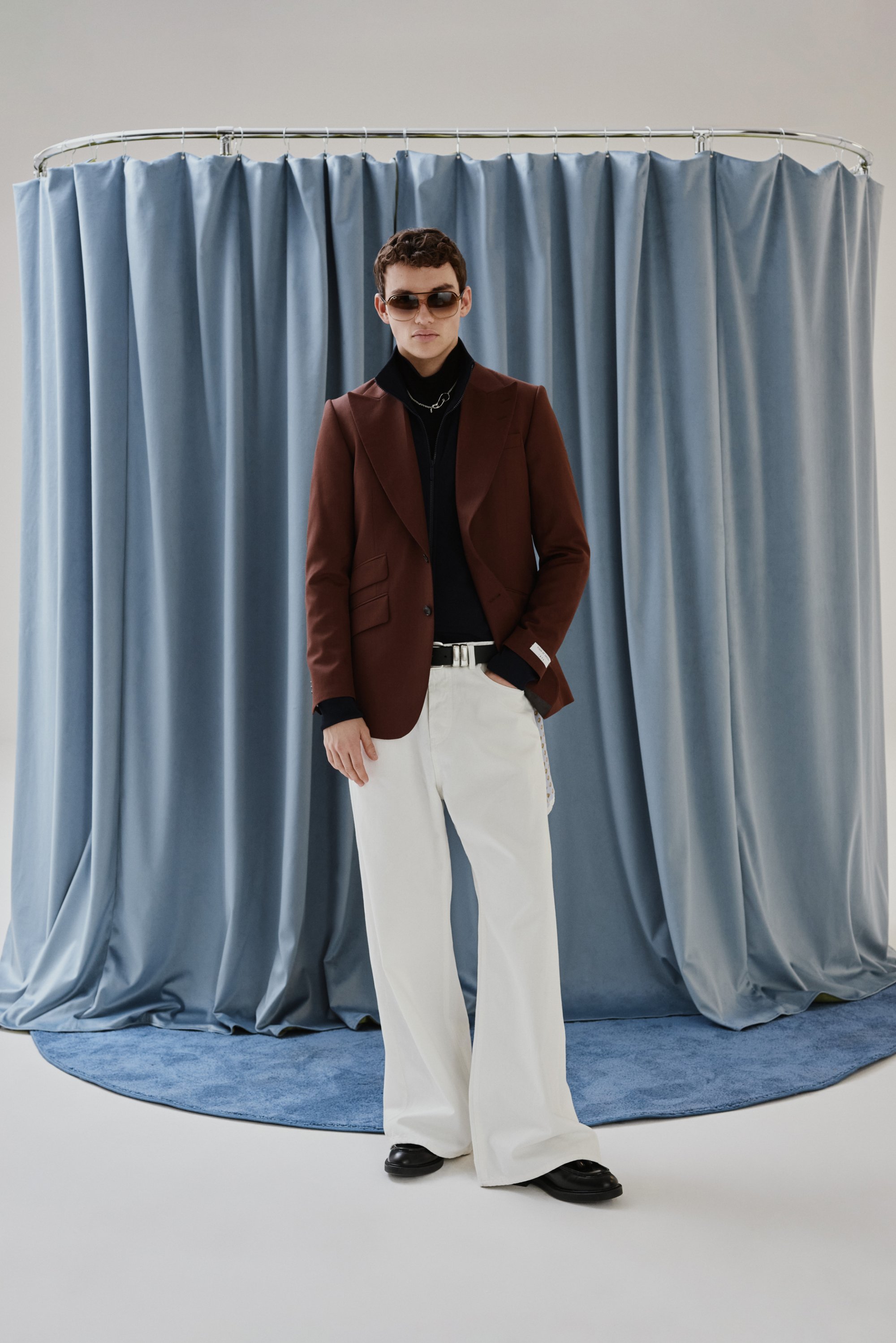 Un modelo con gafas de sol se presenta con confianza en un elegante blazer marrón, cuello alto negro y pantalones blancos, rodeado de suaves cortinas azules.