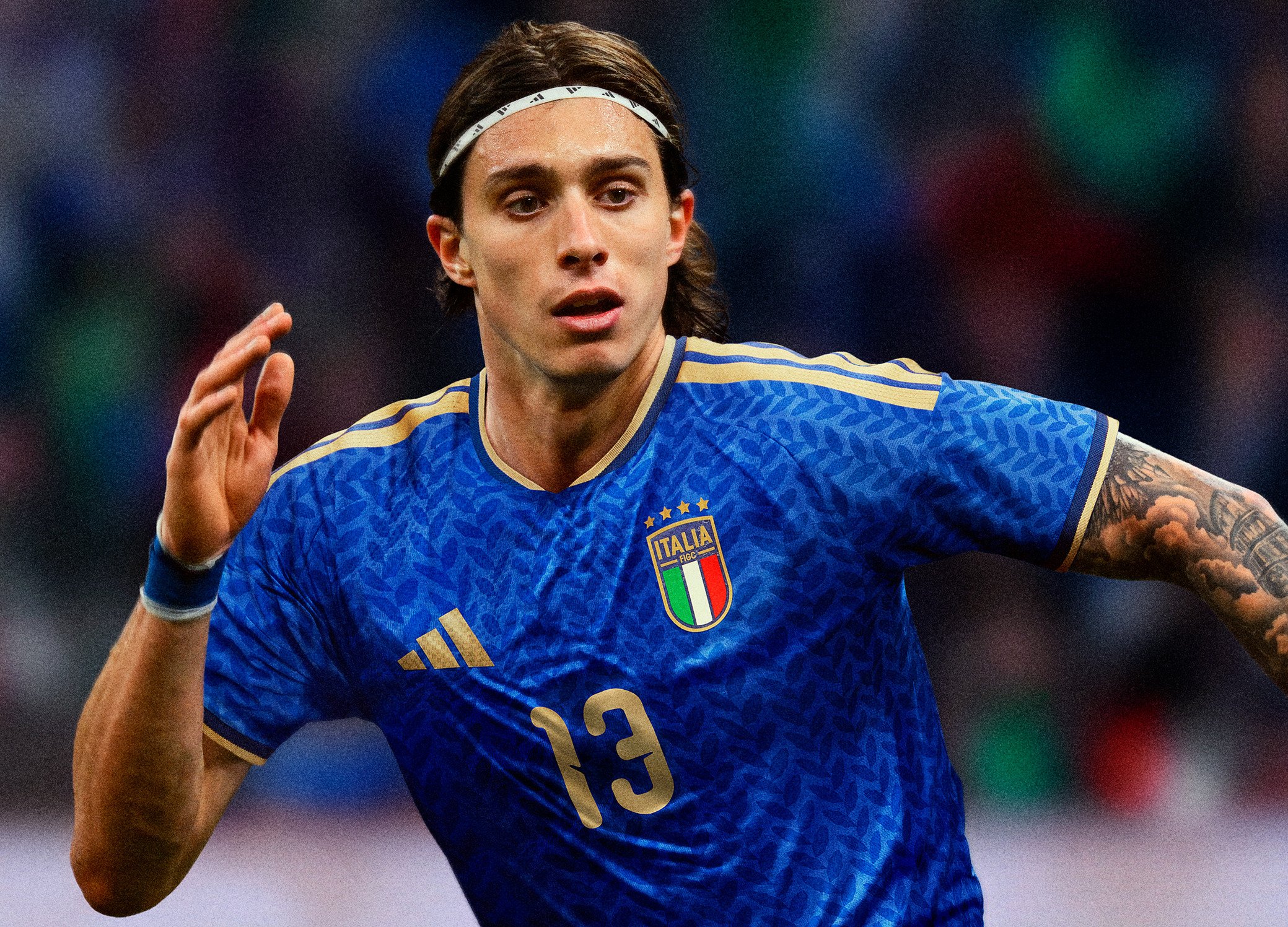 Włoski piłkarz w koszulce z numerem 13 i tatuażami biegnący podczas meczu, ubrany w niebieski strój Adidas z białą opaską na głowie.