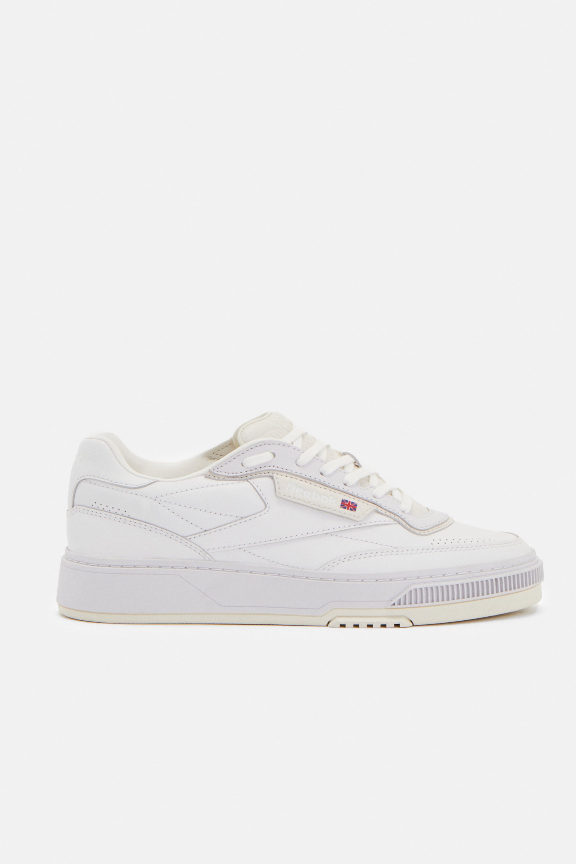 Zapatilla deportiva blanca con superficie lisa, punta redonda y suela texturizada. Las características incluyen cordones y un pequeño logotipo en el lateral.