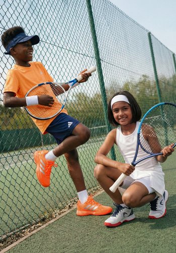 Zwei Kinder auf einem Tennisplatz, die Schläger halten; eines trägt orange Sportkleidung und ein Visier, das andere ein weißes Tennisoutfit mit Stirnband.