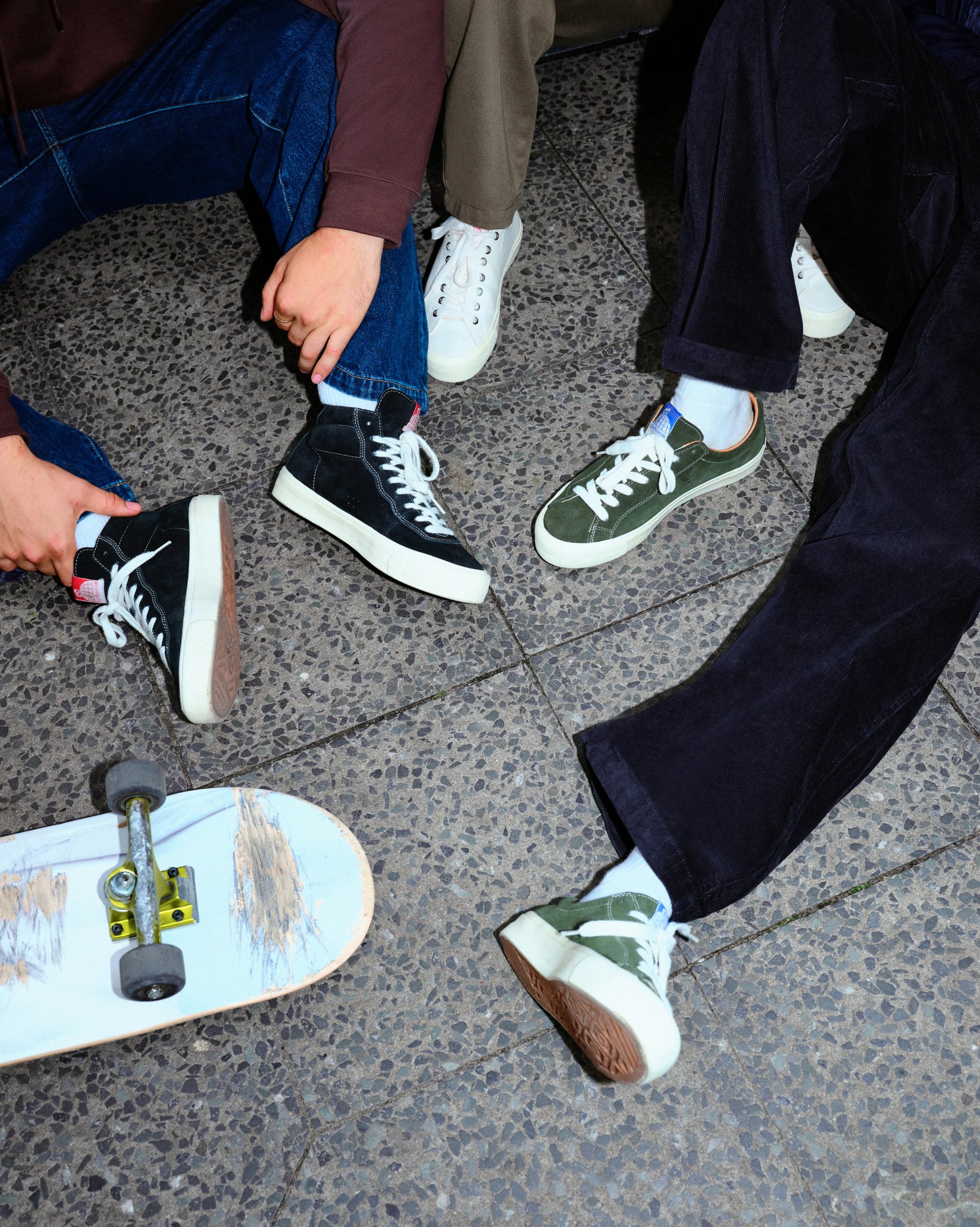 Four pairs of sneakers on a textured pavement, with a worn skateboard nearby. Hands rest on knees, emphasizing casual, urban style.
