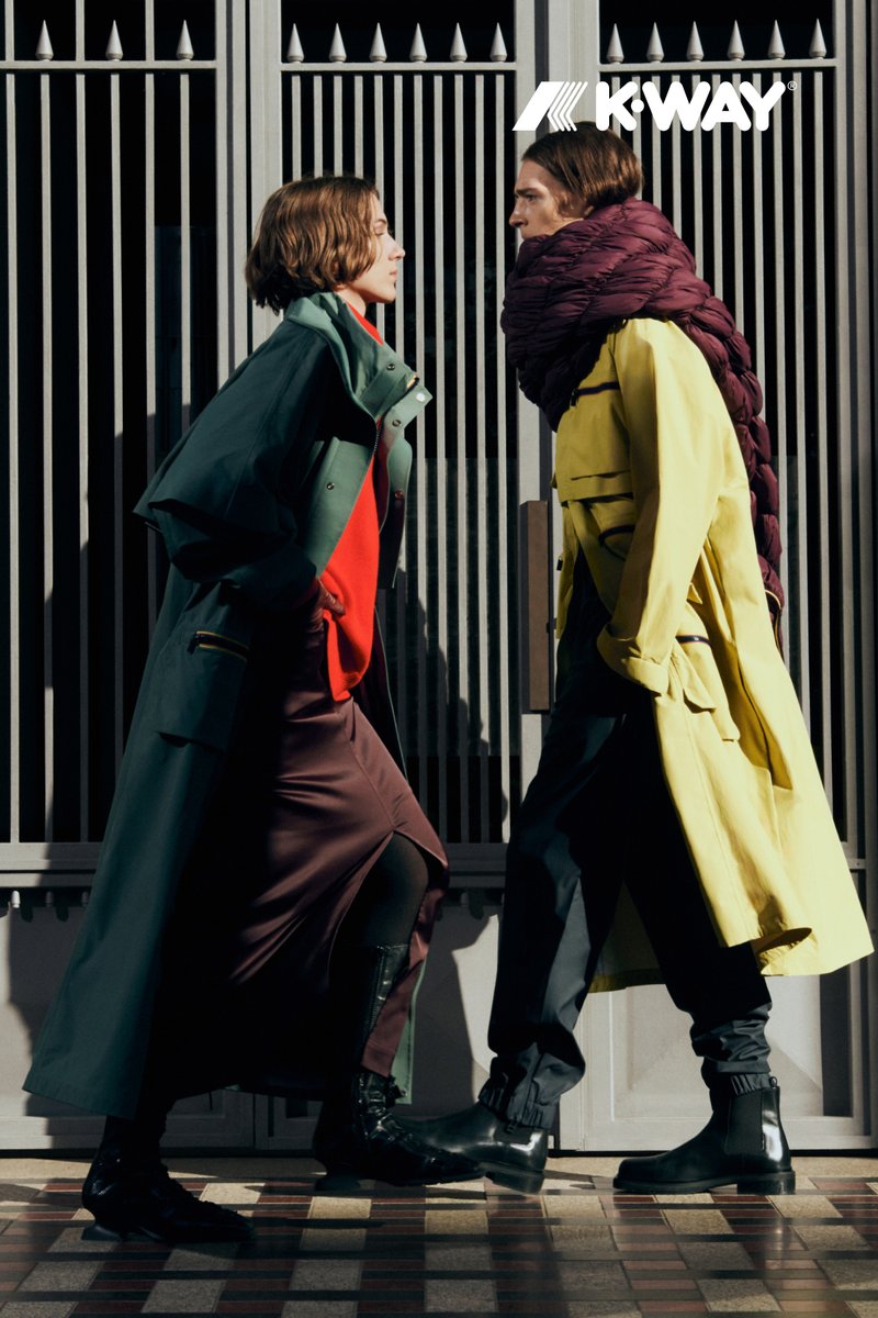 Due modelle camminano rapidamente, indossando un lungo impermeabile giallo e un impermeabile verde sopra un maglione rosso, con pantaloni scuri e stivali neri.