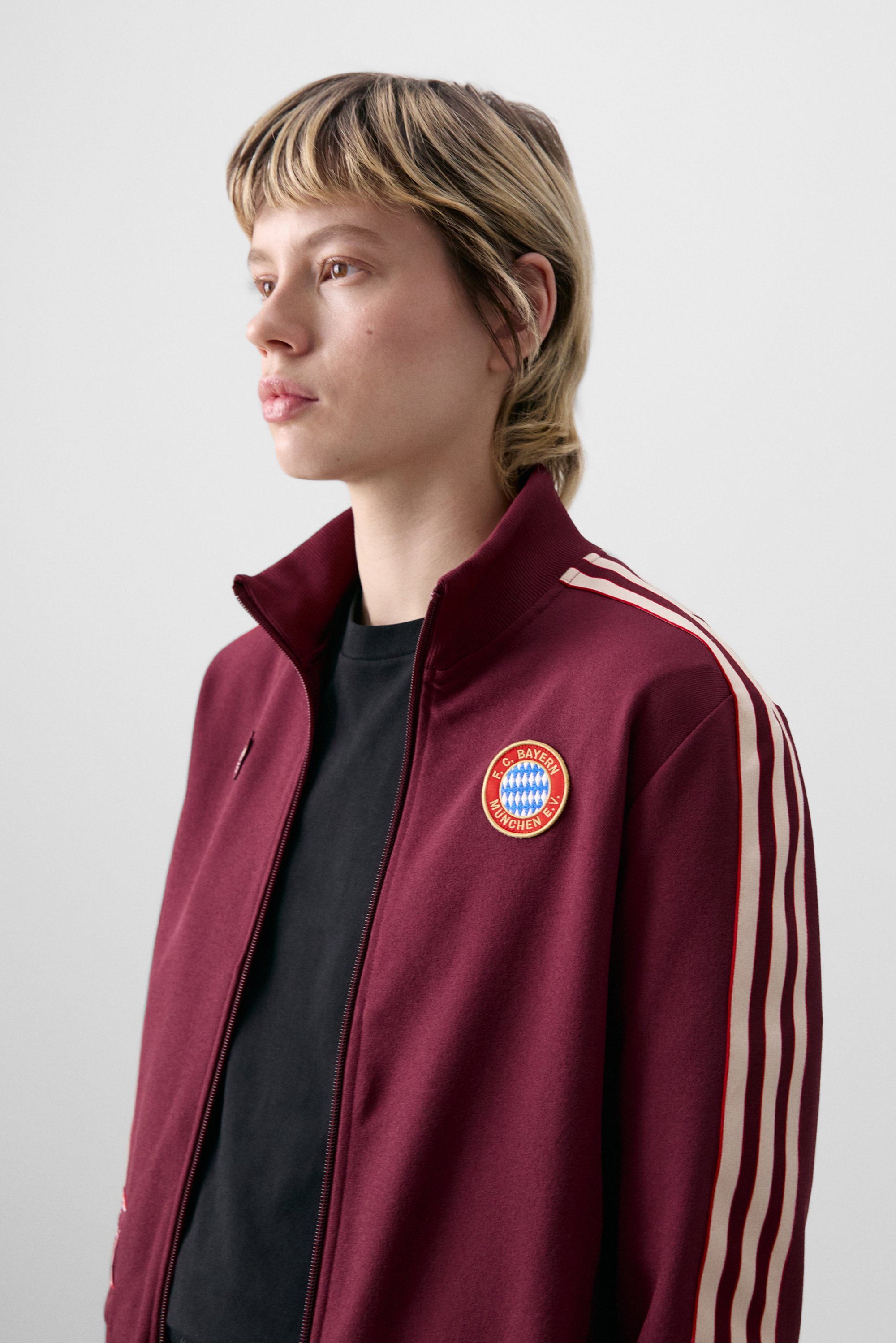 A person with short, tousled hair wears a maroon sports jacket with FC Bayern Munich's logo, black t-shirt underneath, against a neutral background.