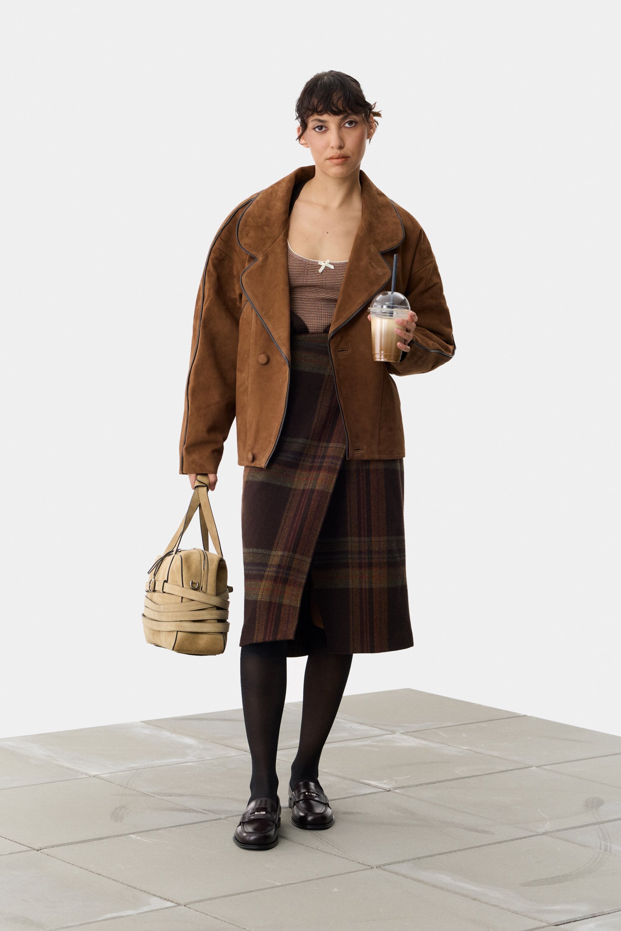 Chaqueta grande de ante marrón, blusa a rayas, falda de cuadros marrón, medias negras, mocasines marrones, bolso beige, sosteniendo una bebida.