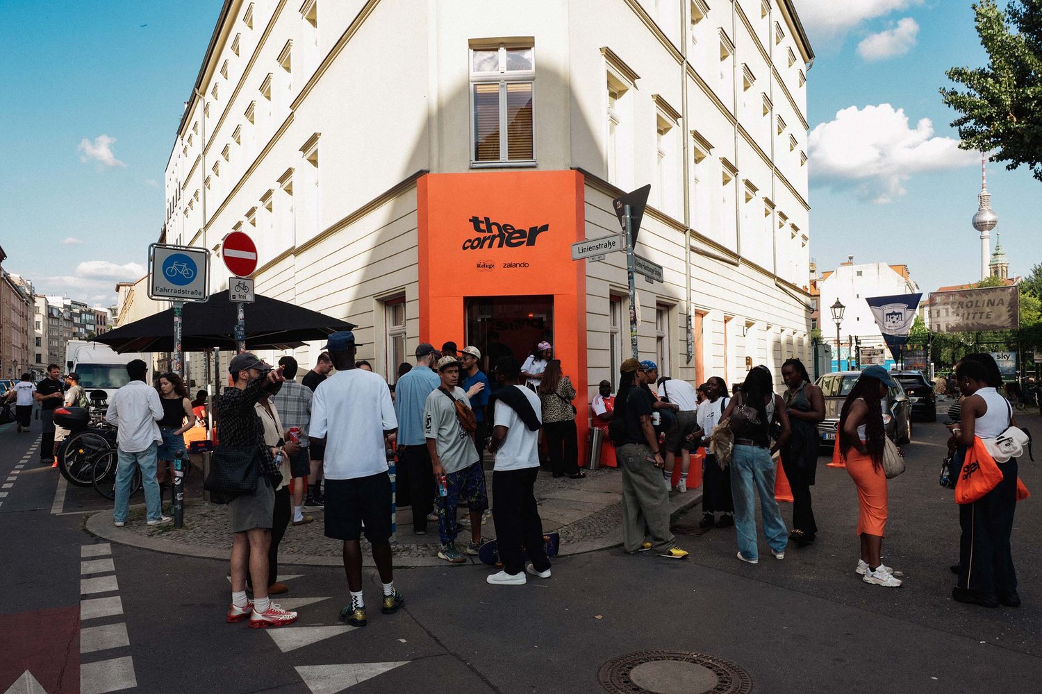 Udsigt til "hjørnet" butik med en orange facade. En mangfoldig gruppe mennesker er samlet udenfor og engageret i samtale. Vejskilte er synlige.