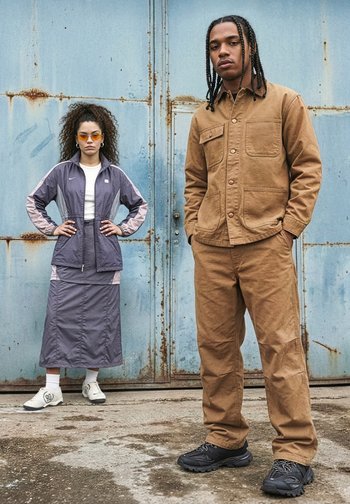 Two young adults stand against a rusty blue metal door; one wears a brown jacket and trousers, the other a purple tracksuit and white trainers.