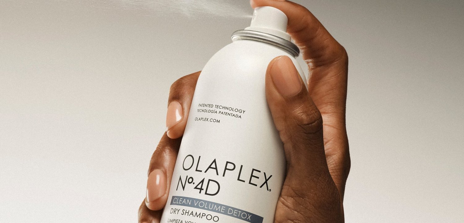 Ręka o ciemnej karnacji trzymająca biały spray do suchego szamponu Olaplex No. 4D clean volume detox na jasnym tle.