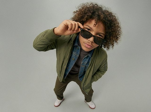 Jeune personne aux cheveux bouclés portant une veste verte et des lunettes de soleil, regardant vers le haut tout en ajustant ses lunettes, debout sur un fond gris uni.