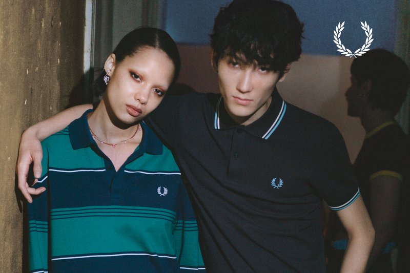 Two young people wearing Fred Perry polo shirts stand close together indoors, one with an arm around the other’s shoulder.