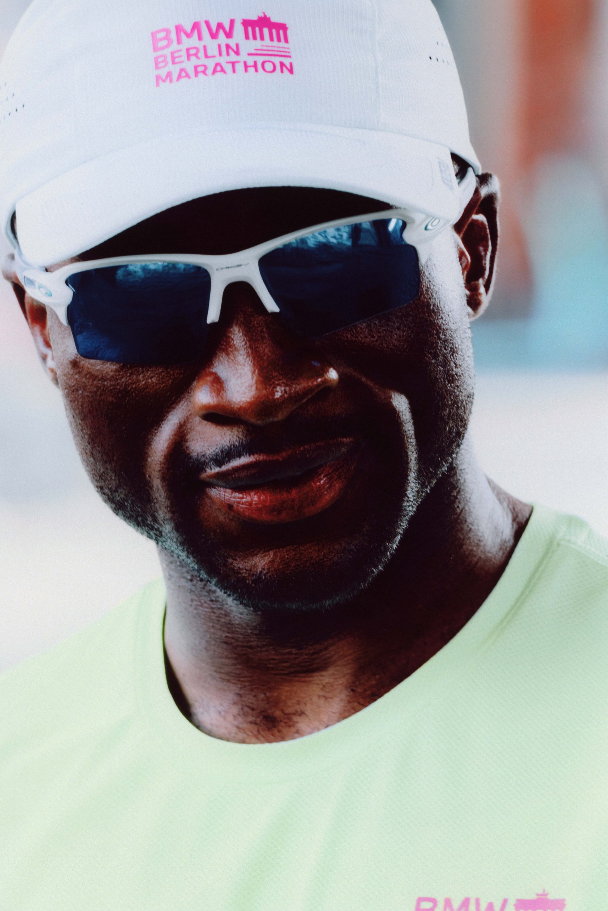 White cap featuring a pink "BMW Berlin Marathon" logo, dark sunglasses, and a light green athletic shirt with a textured fabric pattern.