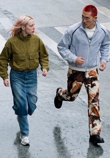 Deux personnes sur un trottoir mouillé ; l'une marche en veste verte et jupe en jean, l'autre court en veste rayée et pantalon à motif cheval.