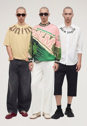 Drie mannen staan naast elkaar in streetwear: oversized beige overhemd met donkere jeans, veelkleurige grafische trui met witte broek, wit overhemd met zwarte cropped broek.