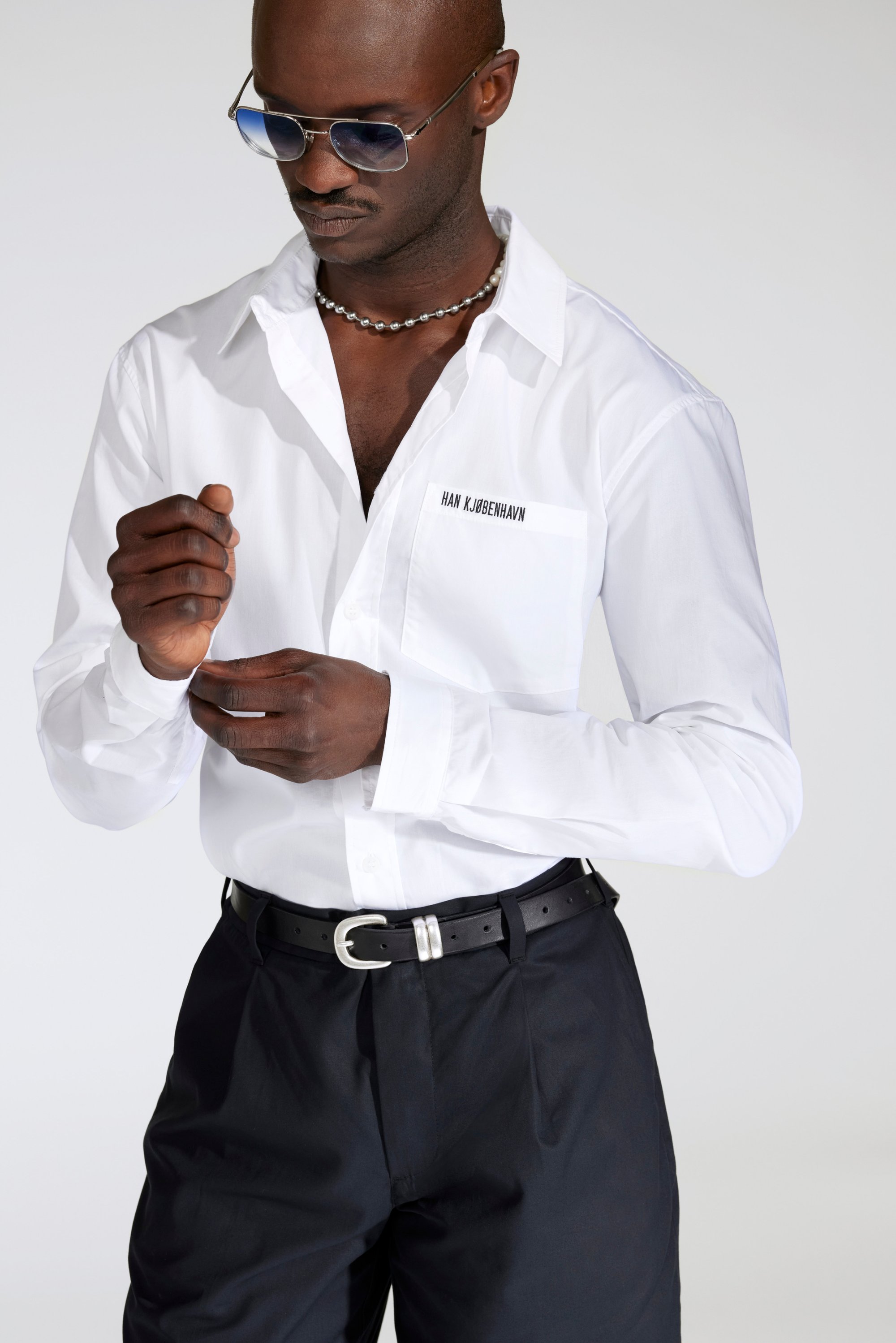 A man with dark skin adjusts his cuff while wearing a white shirt and sunglasses, highlighting a stylish, modern look.