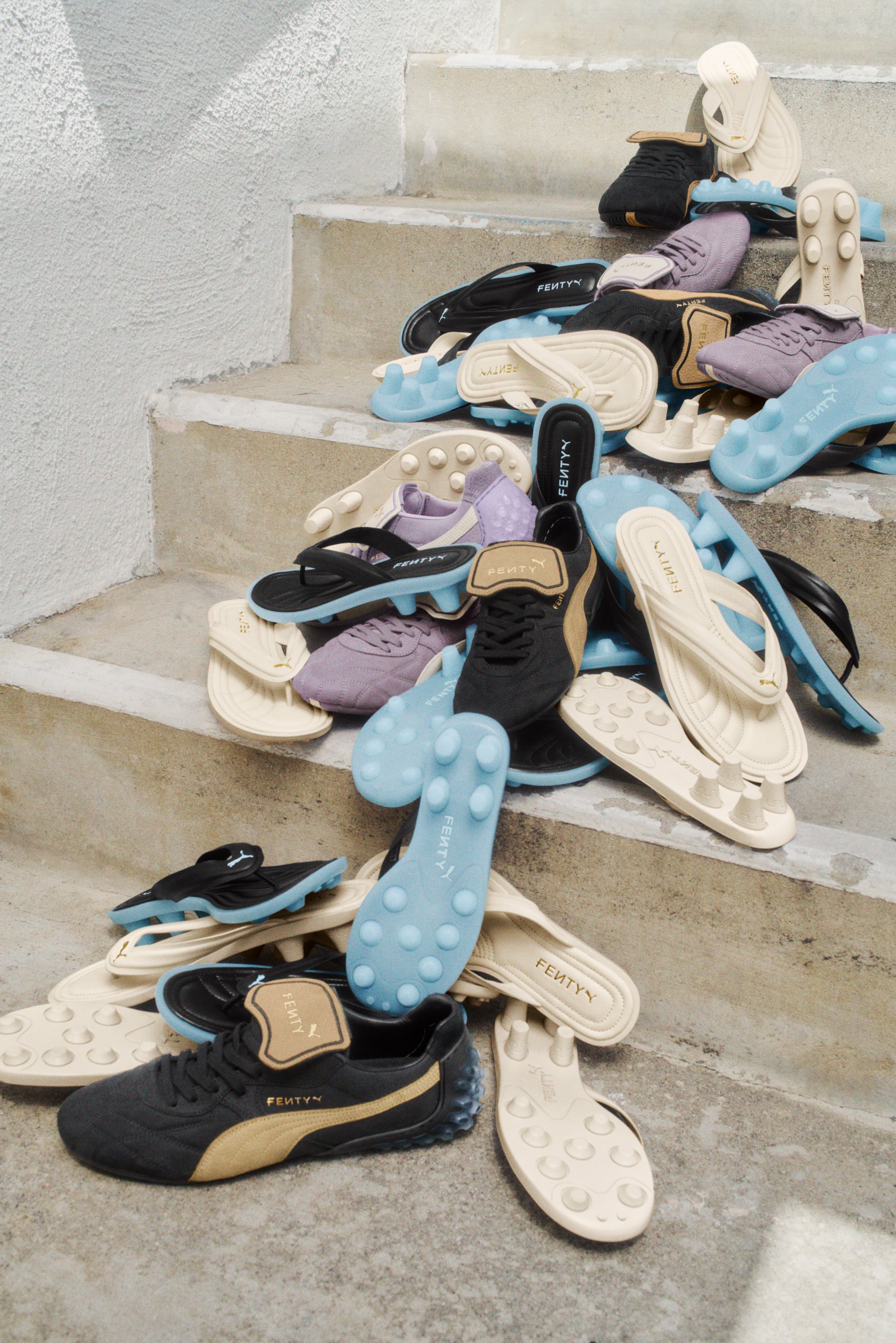 Calzado variado de Fenty en las escaleras: zapatillas negras y beige, chanclas azules y crema, y flip-flops morados, con texturas y diseños diversos.