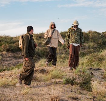 Drie individuen wandelen op een zandpad door hoog gras, gekleed in outdoor kleding. Een vrouw draagt een tas, terwijl de anderen de omgeving verkennen.