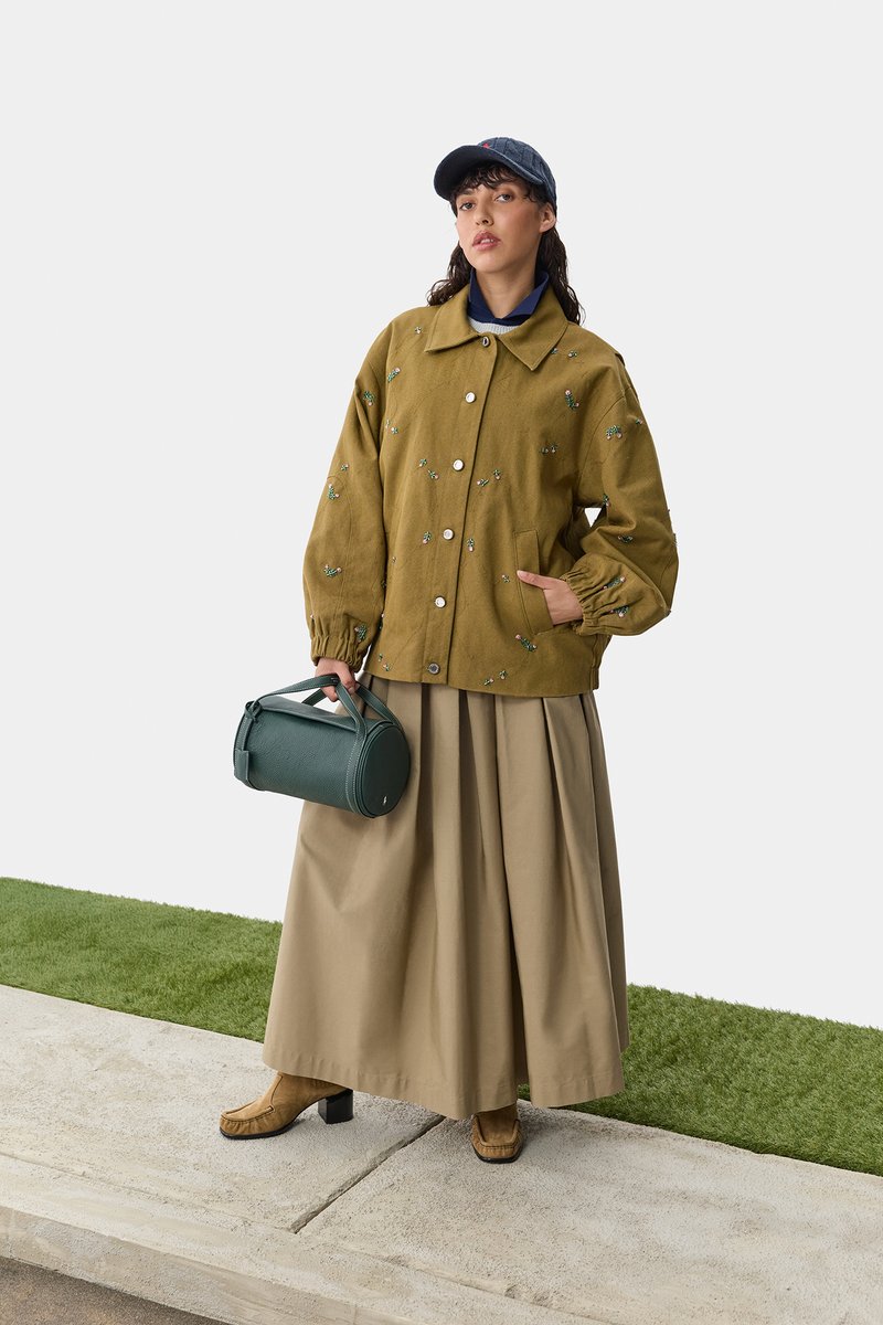 Olivgrön jacka med blommönsterbroderi, beige plisserad kjol, bruna klackboots och en mörkblå keps, håller en grön cylindrisk väska.