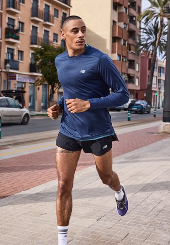 Άνδρας δρομέας με μπλε μπλούζα μακρυμάνικη και μαύρο σορτς τρέχει στο πεζοδρόμιο της πόλης με κτίρια και αυτοκίνητα στο βάθος.