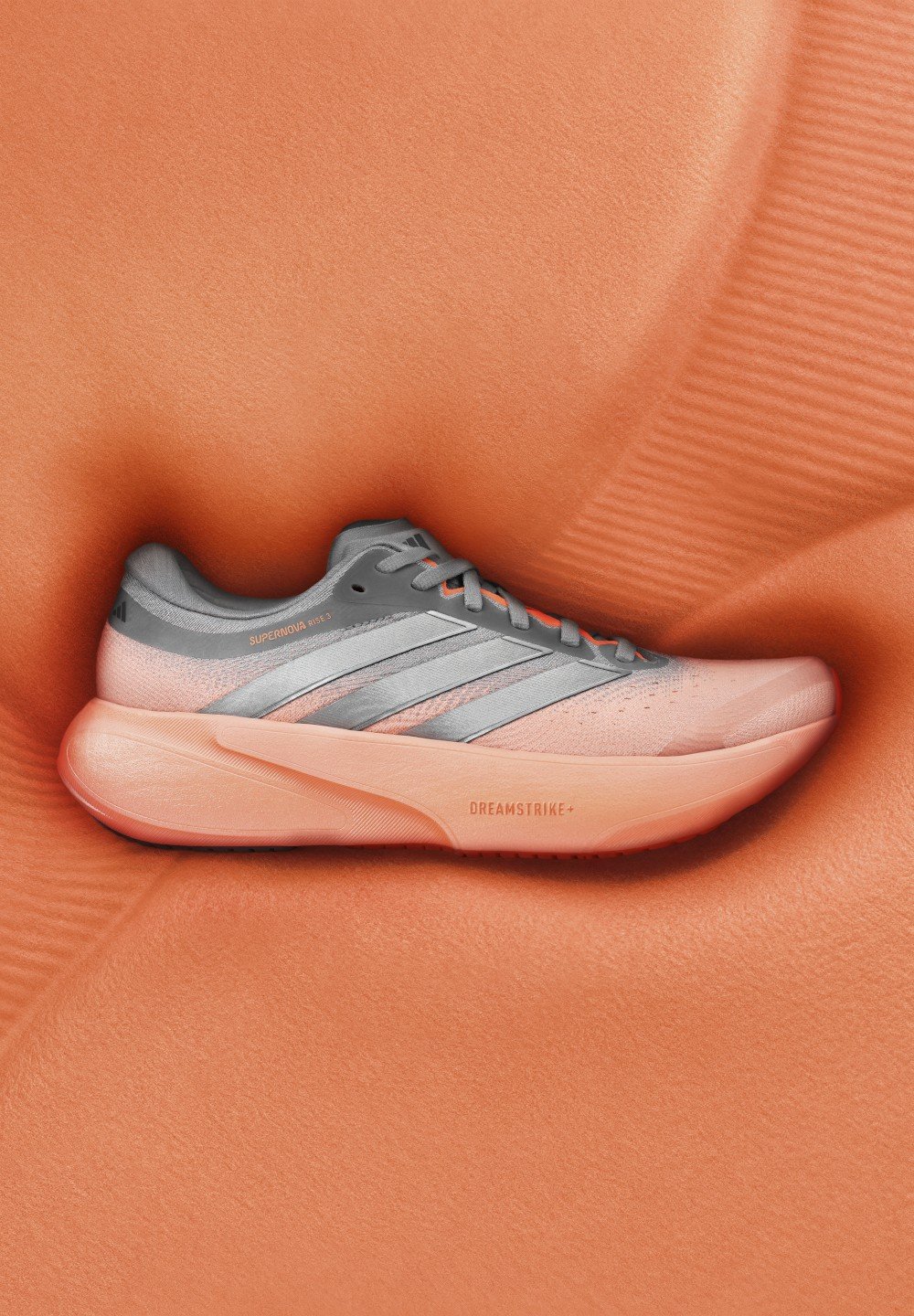 Chaussure de course Adidas grise et corail avec le mot "Supernova" sur la tige et "Dreamstrike+" sur la semelle épaisse, reposant sur une surface corail texturée.