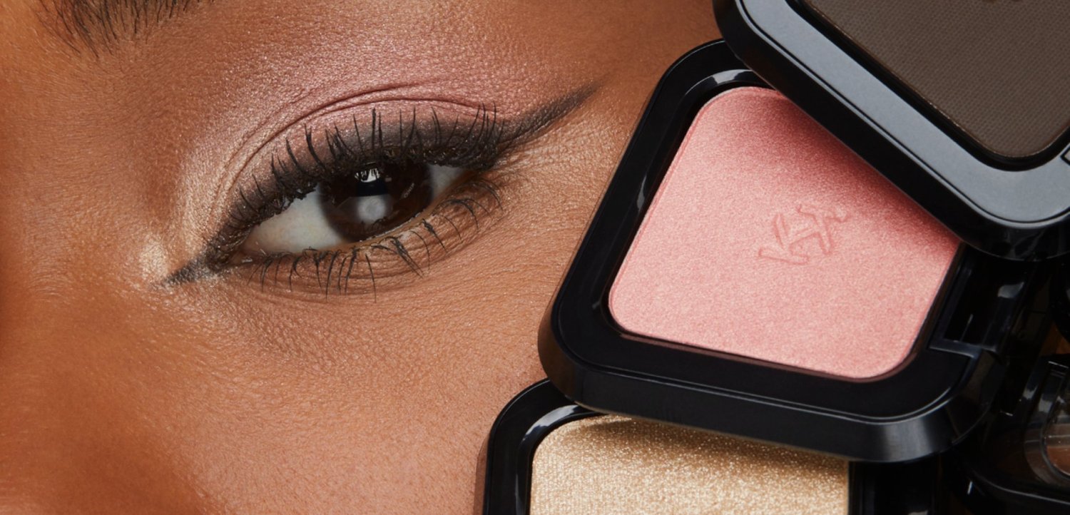 Nahaufnahme eines Auges mit natürlichem Make-up, umgeben von geöffneten schwarzen Kompaktgehäusen mit rosa, braunen und goldenen Pudern.