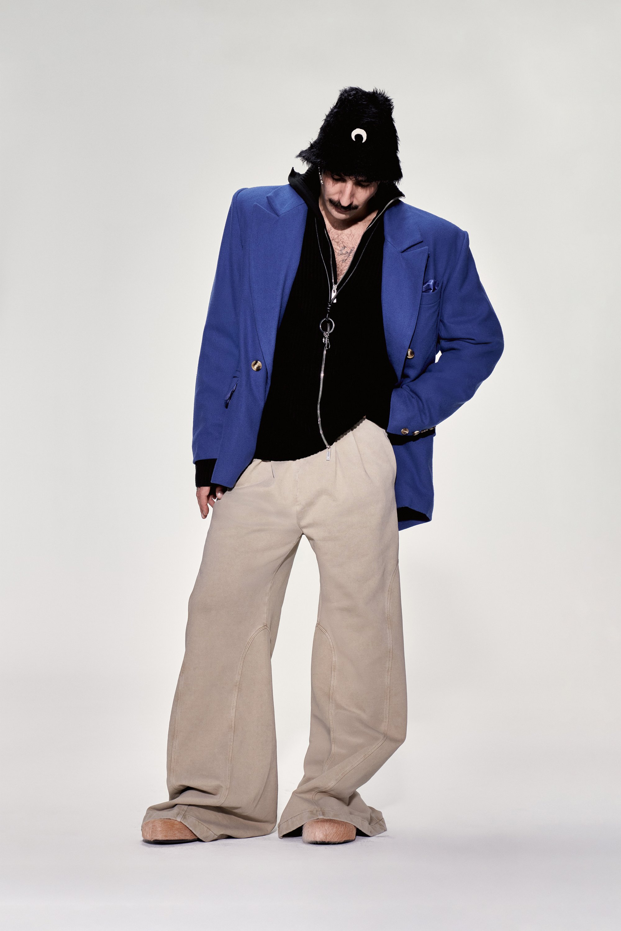 Un hombre se encuentra con un atuendo holgado, llevando un blazer azul sobre una camisa negra, pantalones beige y un sombrero negro de piel sintética, mirando hacia abajo con expresión pensativa.