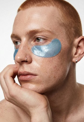 Persoon met kortgeschoren rood haar, die blauwe gelpleisters onder de ogen draagt, met de kin op de hand steunend en nadenkend naar de zijkant kijkt.