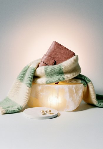 Brūna ādas maks ietīts zaļi-kreminā svītraina adīta šallē, kas atrodas uz caurspīdīga akmens bloka, netālu baltā traukā ir pērļu auskari.