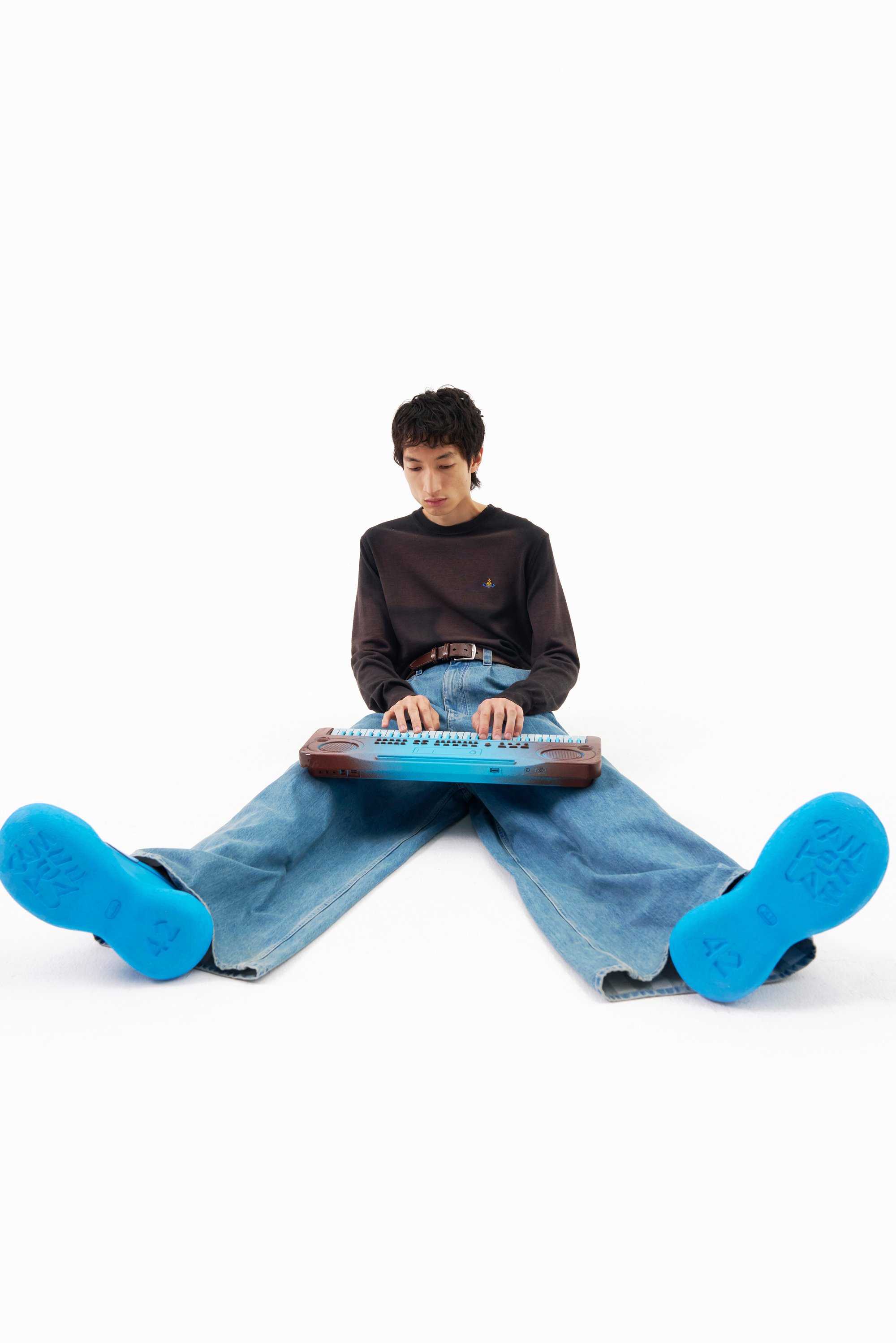 Una persona se sienta con las piernas extendidas sobre un fondo blanco, tocando un teclado musical azul, vistiendo una camisa oscura y pantalones de mezclilla grandes.