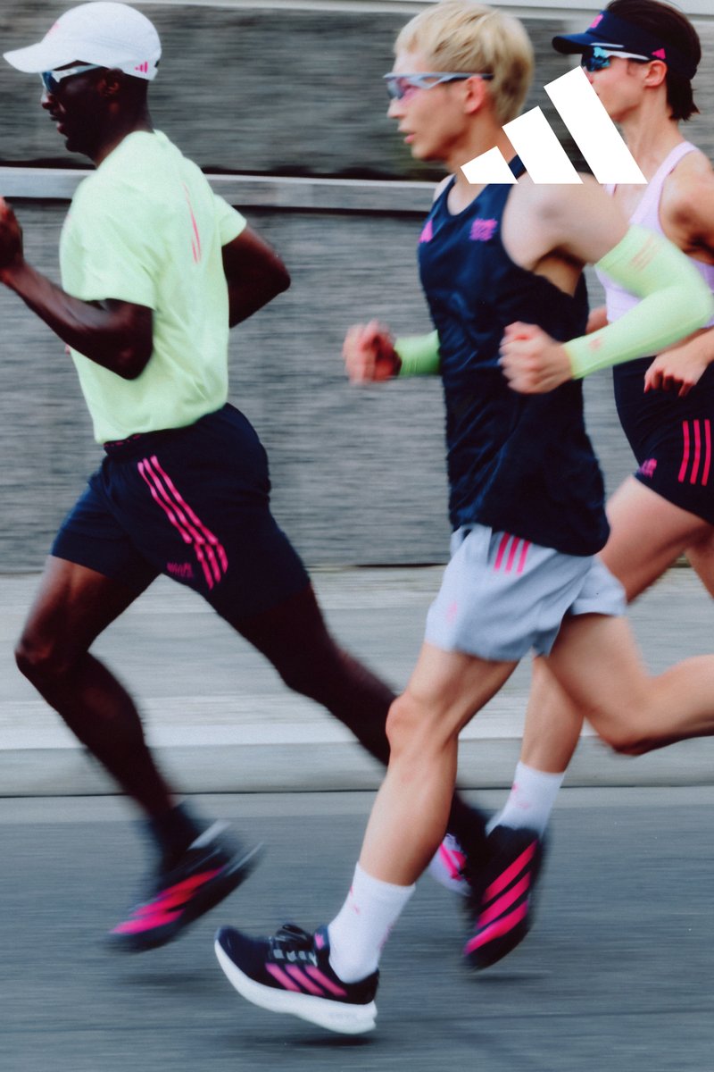 Drie hardlopers in sportkleding en roze gestreepte schoenen rennend op een stadsstraat met een betonnen muur op de achtergrond.