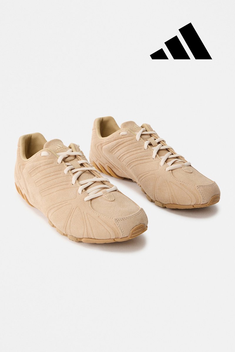 Chaussures de sport beiges en daim avec des panneaux texturés, des lacets blancs et des semelles en caoutchouc, arborant un logo à trois bandes noir.