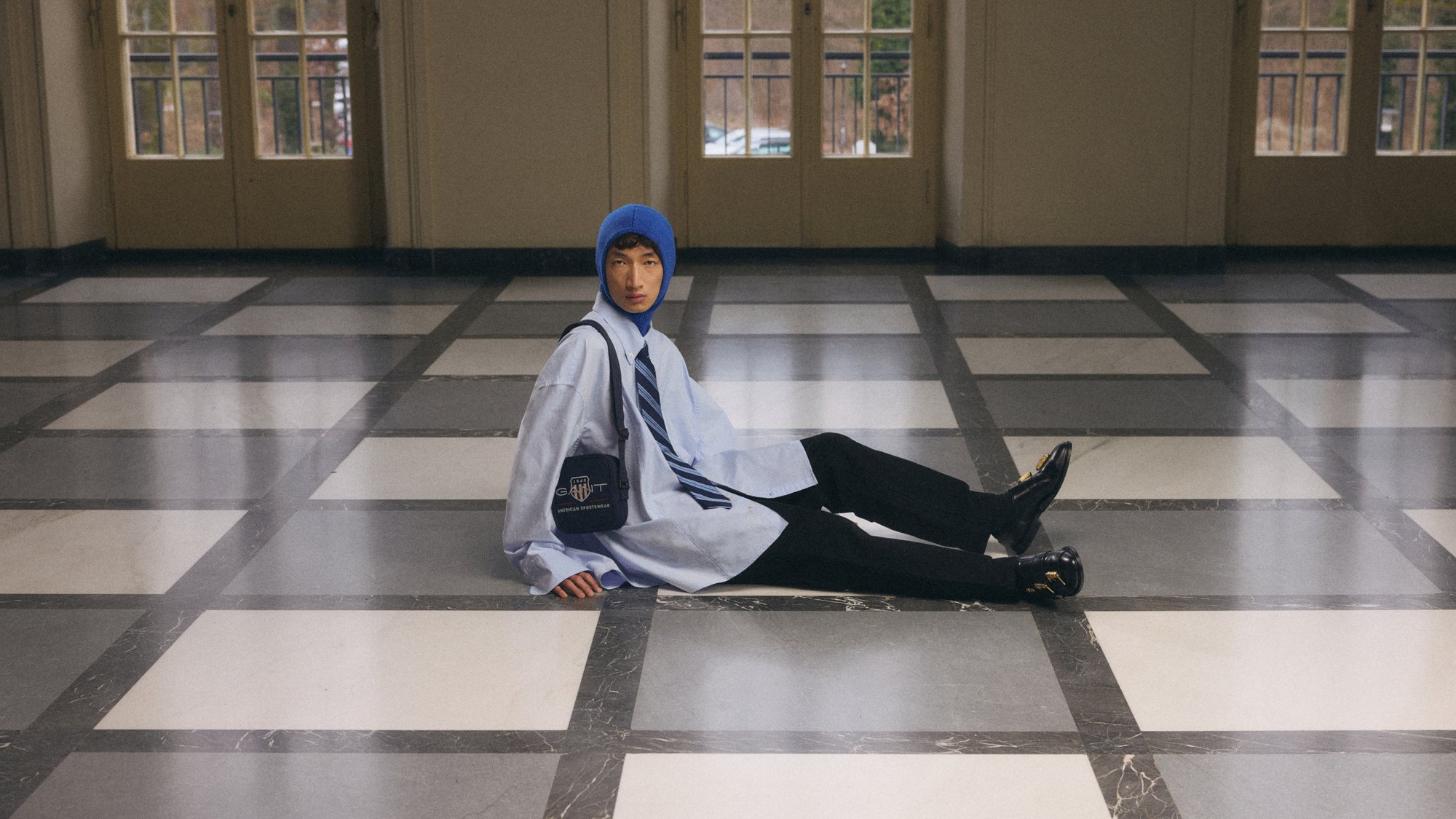 En person sitter på ett rutigt golv, iklädd en blå mössa och en överstor skjorta med en slips, och håller en liten väska. Stora fönster finns i bakgrunden.