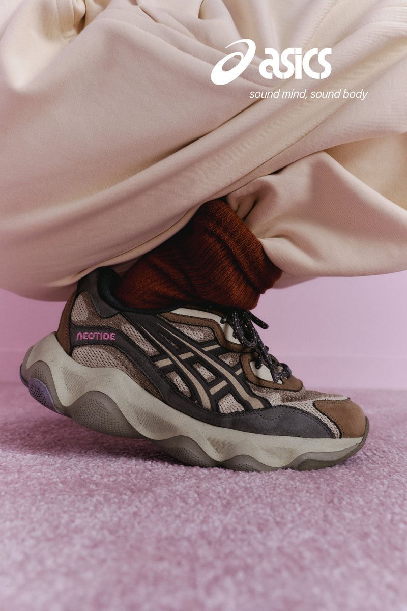 Sneaker ASICS Neoride beige e marrone indossate con calzini color ruggine e pantaloni beige su tappeto rosa, con logo ASICS e slogan visibili.