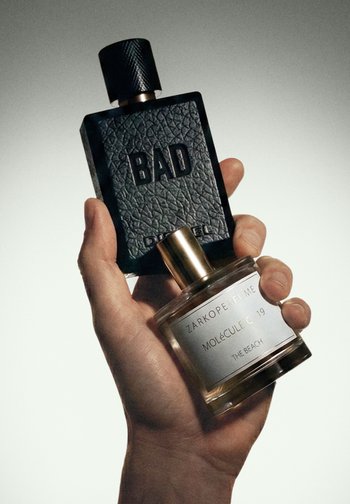Hånd holder to parfumeflasker: en sort med "BAD" prægning, og en klar med mærkatet "Zarkoperfume Molécule C 19 The Beach."
