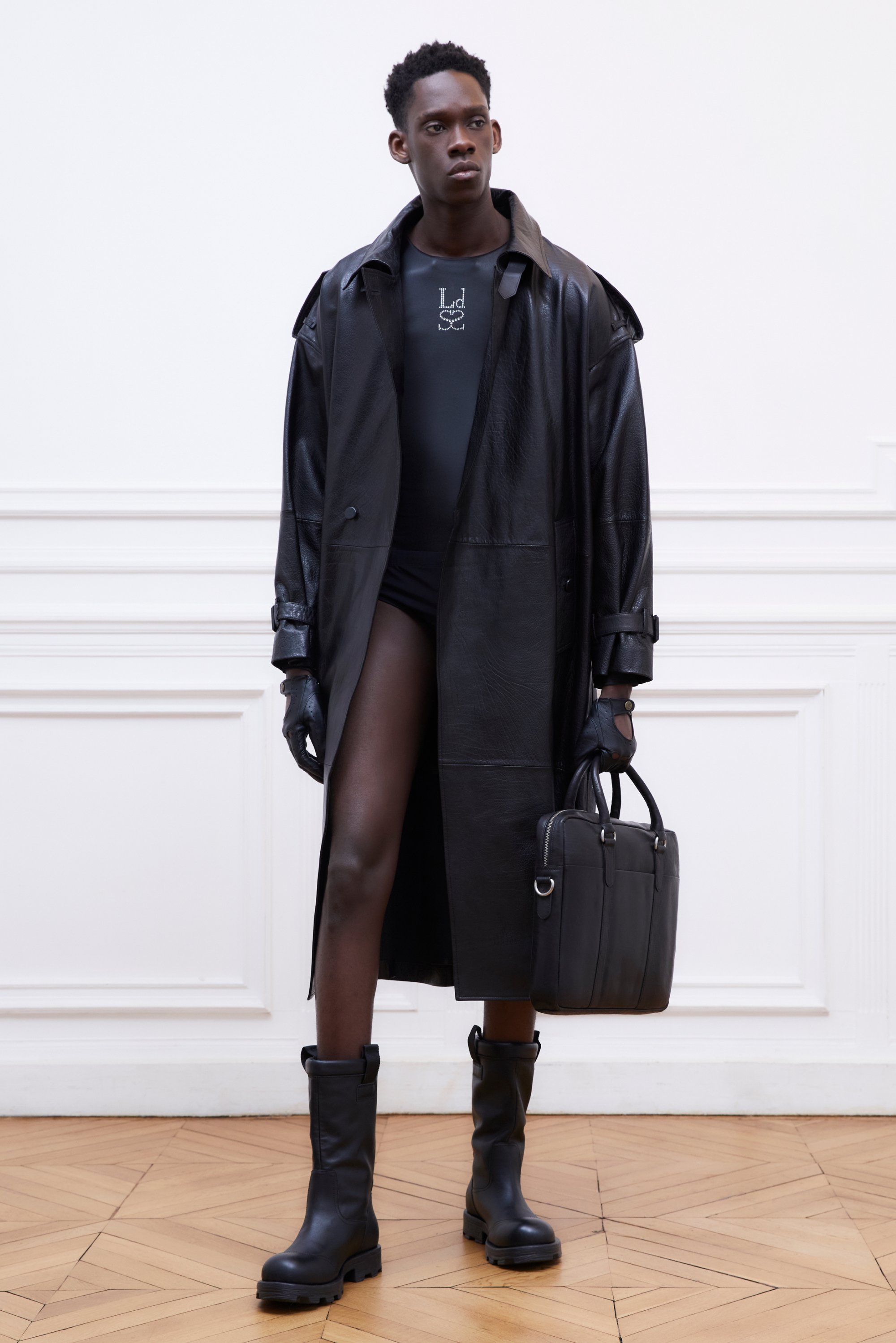 Una modella posa in una stanza minimalista, indossando un trench coat in pelle nera, guanti, un bodysuit nero aderente e stivali neri fino al ginocchio, mentre tiene una borsa.