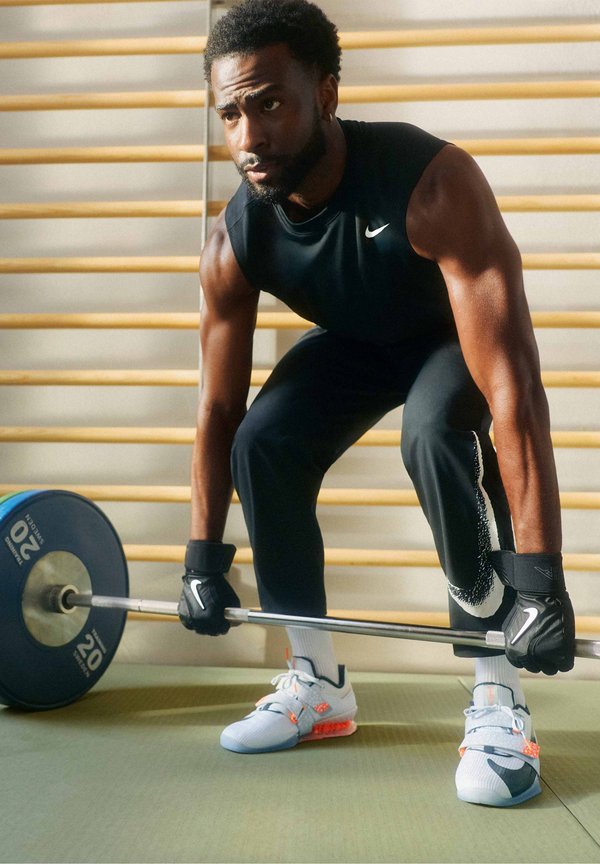 Man wearing black athletic gear and gloves prepares to lift a barbell with 20 kg weights in a gym with wooden wall bars.