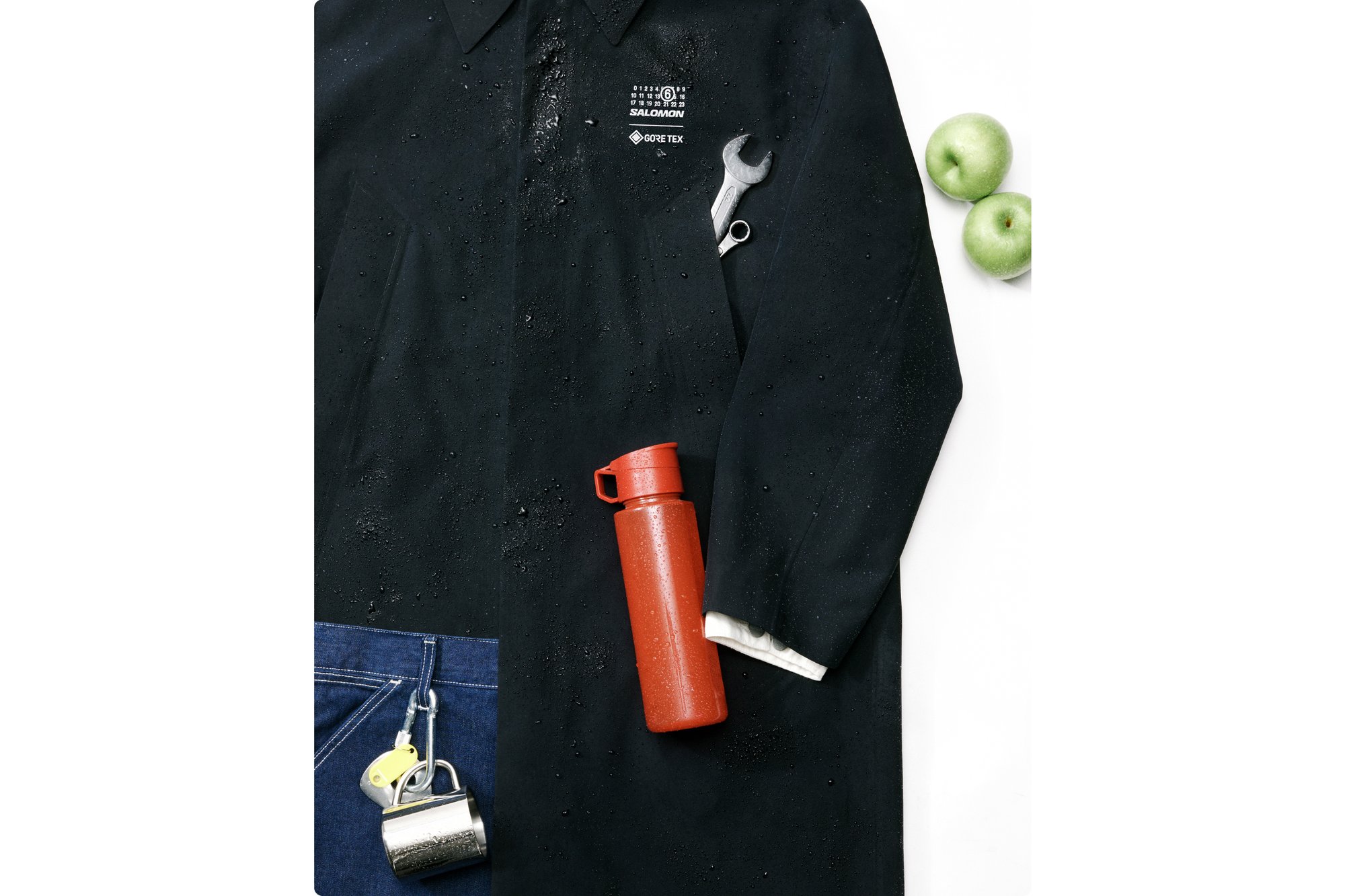 Een zwarte waterdichte jas met druppels, een zilveren moersleutel in de zak, een rode waterfles, groene appels en een hangslot op denim.
