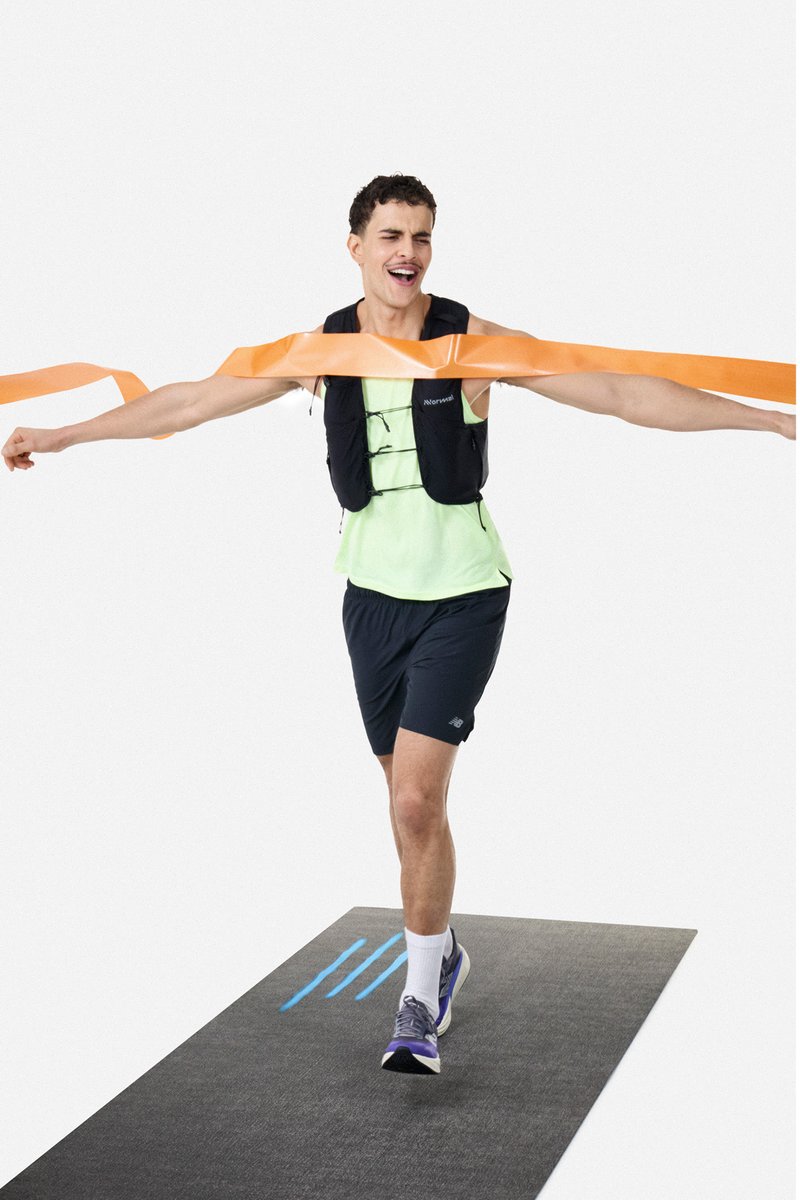 Jonge mannelijke hardloper steekt oranje finishlijn over met uitgestrekte armen, draagt zwarte korte broek, neon top, verzwaarde vest en hardloopschoenen op de baan.