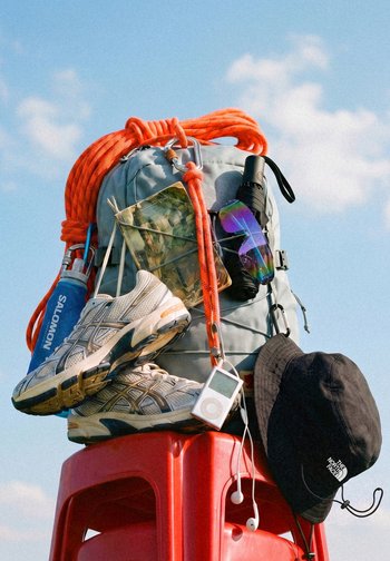 Rucksack mit orangefarbenem Kletterseil, Wanderschuhen, Wasserflasche, gefalteter Karte, Sonnenbrille, Taschenlampe, schwarzer Kappe und Musikplayer auf rotem Hocker im Freien.
