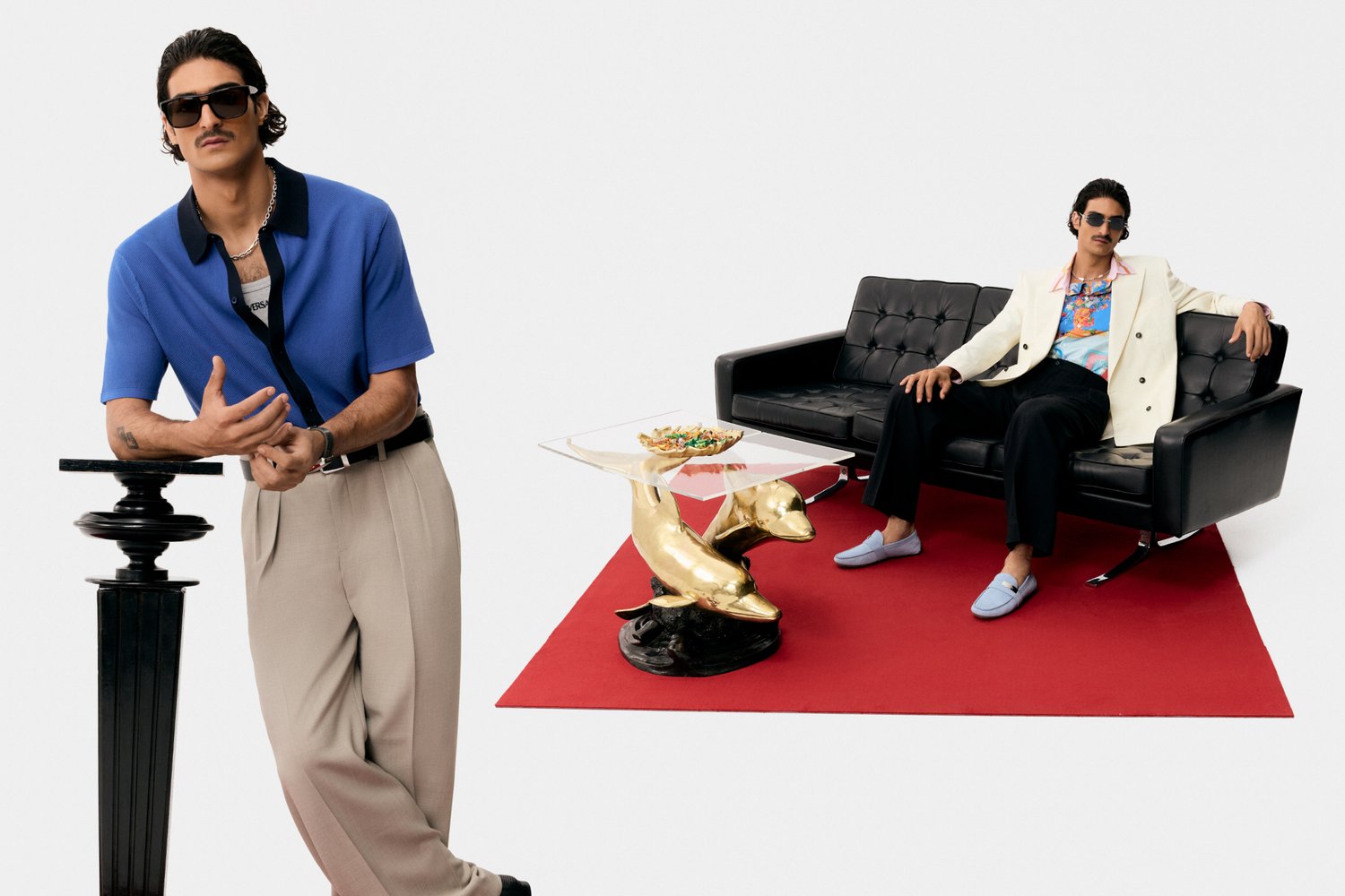 Two stylish men pose in a modern setting. One leans against a decorative column, while the other relaxes on a black sofa, accessorized with funky decor.