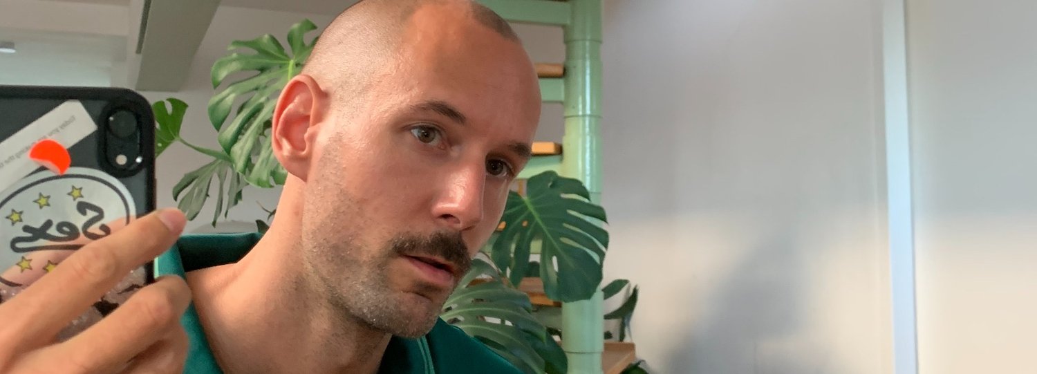 Portret van een persoon met een kaal hoofd en snor, gekleed in een teal shirt, met een smartphone met een decoratieve hoes in de hand, op de achtergrond planten.