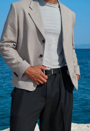 Ljusgrå blazer med tre knappar, bärs över en grå t-shirt. Svarta byxor och bälte fulländar outfiten, mot en blå vattenbakgrund.