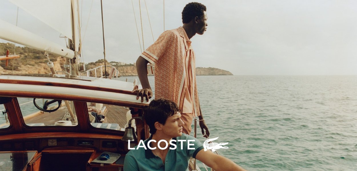 Due persone su una barca, una con una camicia leggera a righe rosa e l'altra con una camicia teal uniforme, sullo sfondo di un paesaggio costiero marino.