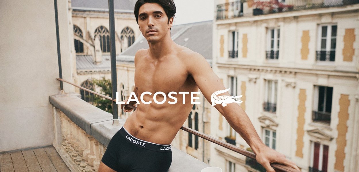 Neri boxer in cotone con una cintura che presenta il logo Lacoste, indossati da un modello in piedi su un balcone con uno sfondo urbano.
