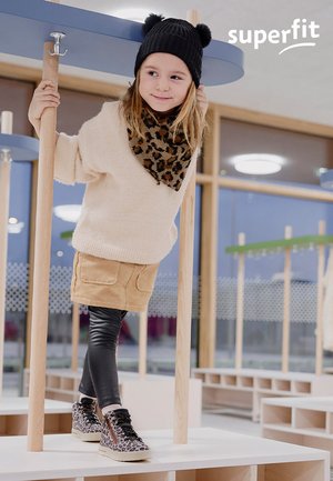 Junges Mädchen mit Leopardenmuster-Schuhen und -Schal, beiger Rock und schwarzem Hut, das drinnen auf Holzpollern balanciert, mit "superfit"-Logo.