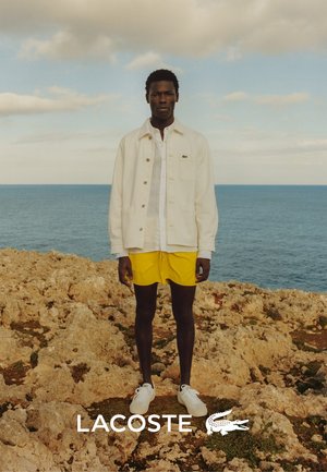 Giacca di jeans bianca sopra una camicia bianca, pantaloni corti gialli e sneakers bianche, in piedi su un terreno roccioso con un mare blu sullo sfondo.