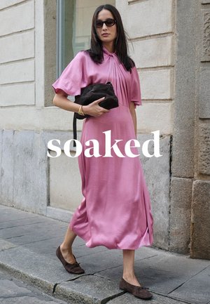 Woman wearing a pink midi dress and sunglasses, carrying a black clutch, walking on a city sidewalk next to a stone building.