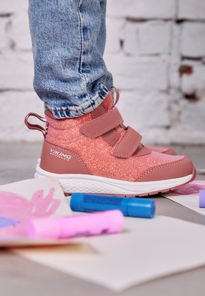 Barn iført pink glitrende viking vandtætte sko med velcro-stropper, stående på gulvet nær farverige tuscher og malet papir.