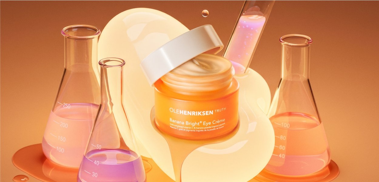 Un pot orange de la crème pour les yeux Banana Bright d'Ole Henriksen avec un couvercle blanc, entouré de béchers en verre et d'une goutte jaune brillante, sur un fond dégradé.